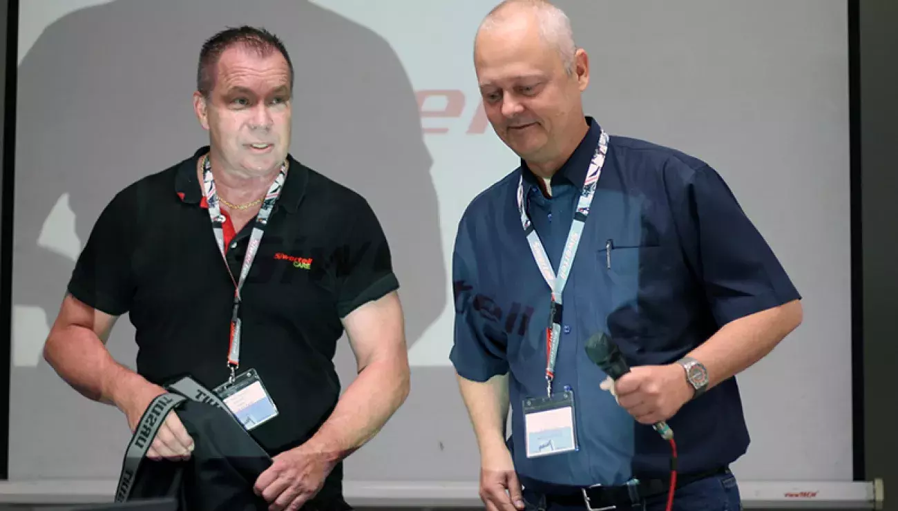 two men in front of a white screen, presenting with microphones